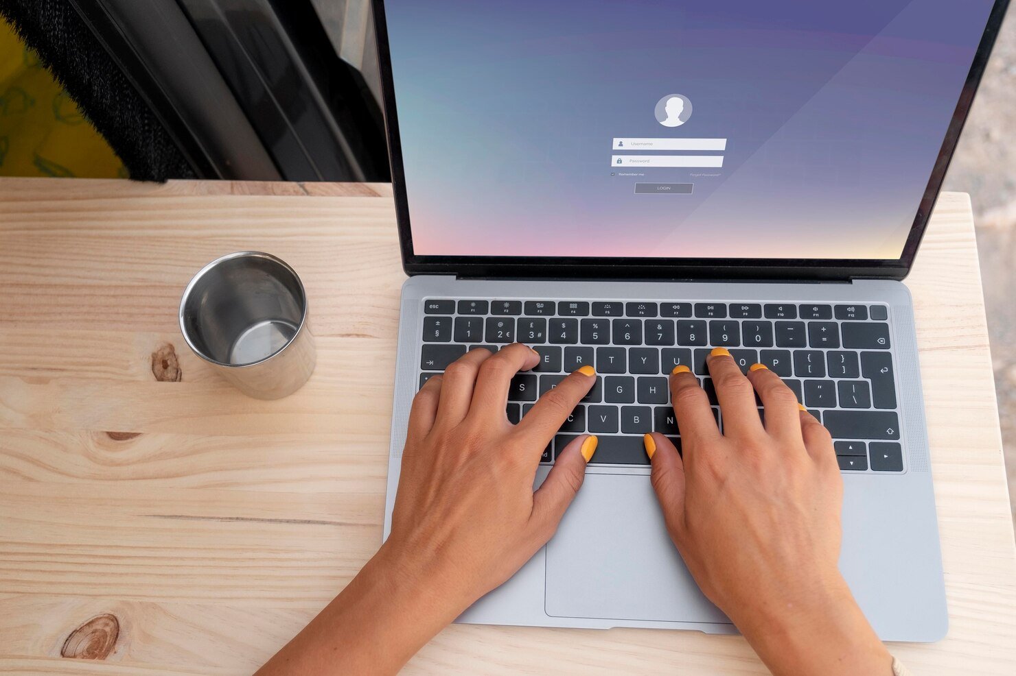 Hands with yellow-painted nails type on a laptop with a login screen displayed. A metal cup sits beside it on a wooden desk.