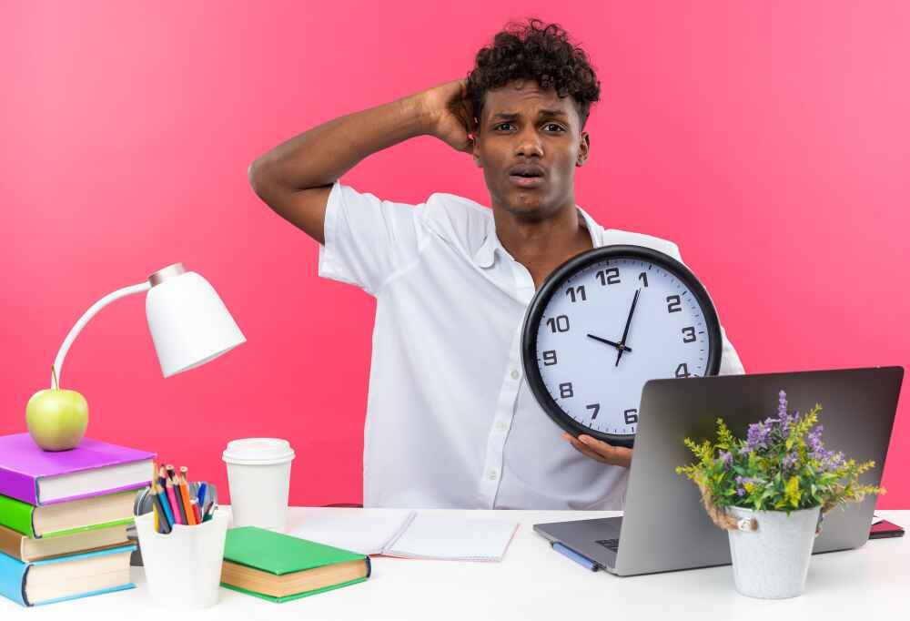 A student sitting behind a desk and managing their time Young man in a white shirt looks stressed, holding a clock at a desk with books, a laptop, and a lamp. The background is pink, suggesting urgency.