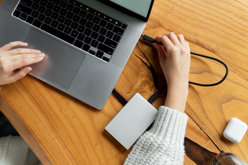 A person in a white sweater is using a laptop on a wooden table. They are connecting a gray external hard drive. Nearby are a pair of wireless earbuds.